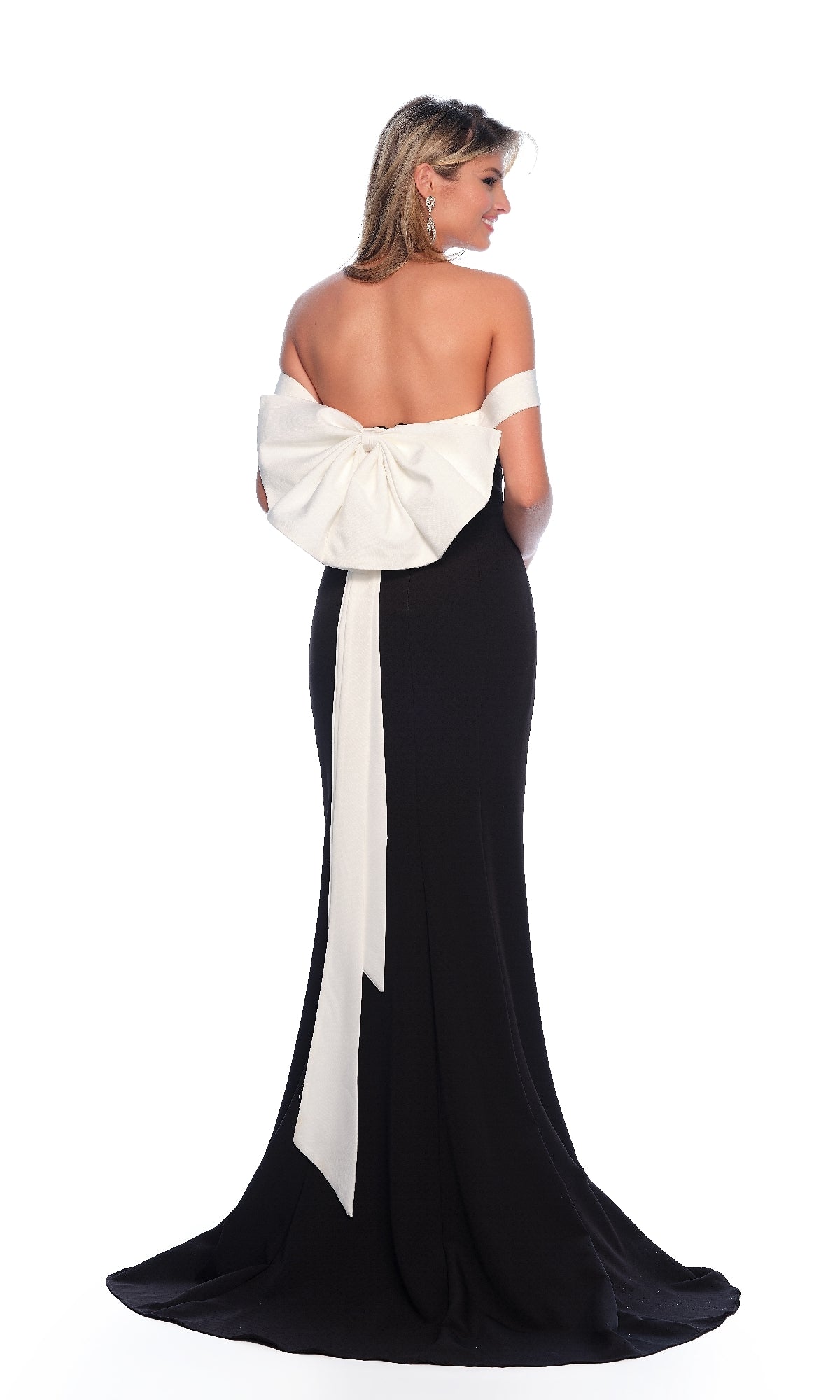 A woman stands against a white background wearing the Dave & Johnny 11786 long black prom dress, an off-the-shoulder floor-length gown with a large white bow and sash on the back.